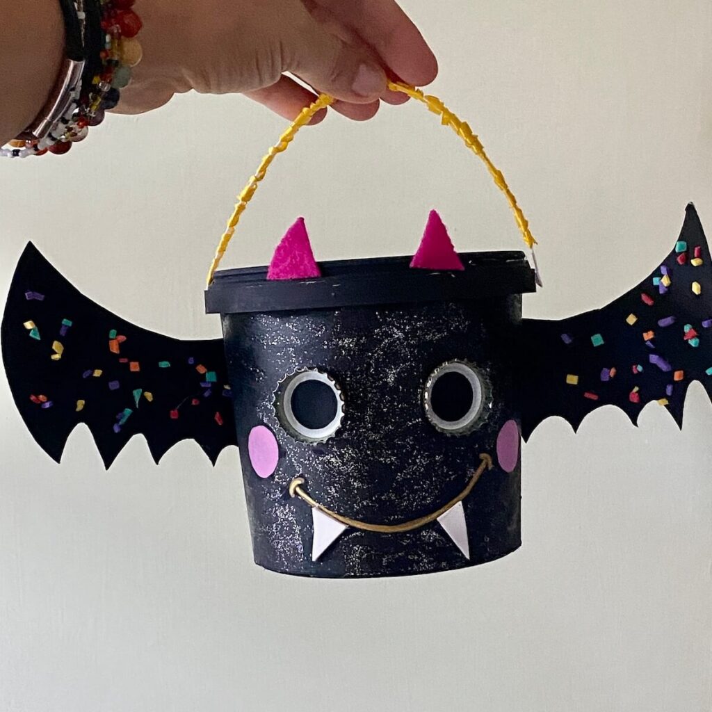 Geniale Halloween Deko basteln: 5 nachhaltige & einfache Ideen 5 Schwarzer Upcycling Joghurteimer als Fledermaus mit Flügeln aus Pappe und Kronkorken Augen für Halloween.